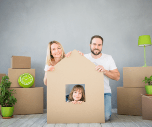 a family of three sit together surrounded by boxes. their child is in the middle inside a home made of cardboard.