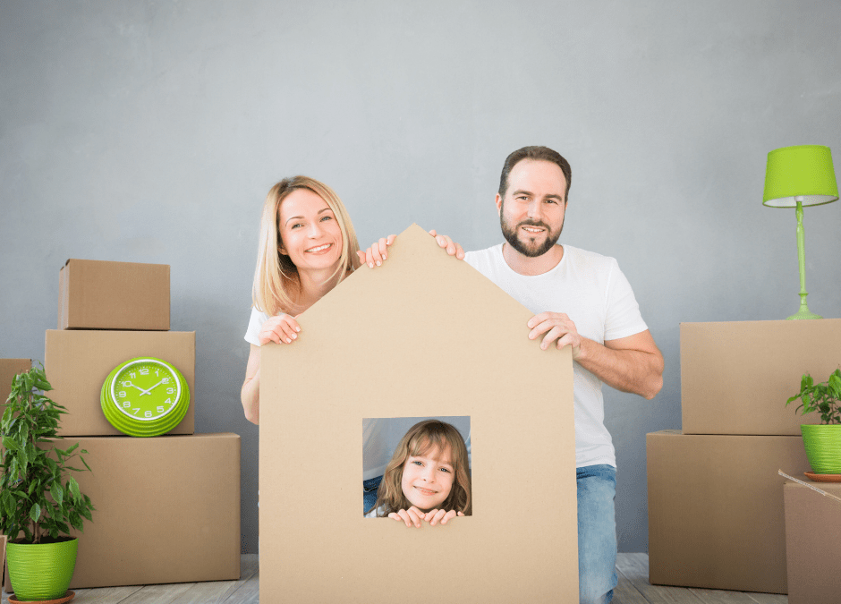 a family of three sit together surrounded by boxes. their child is in the middle inside a home made of cardboard.